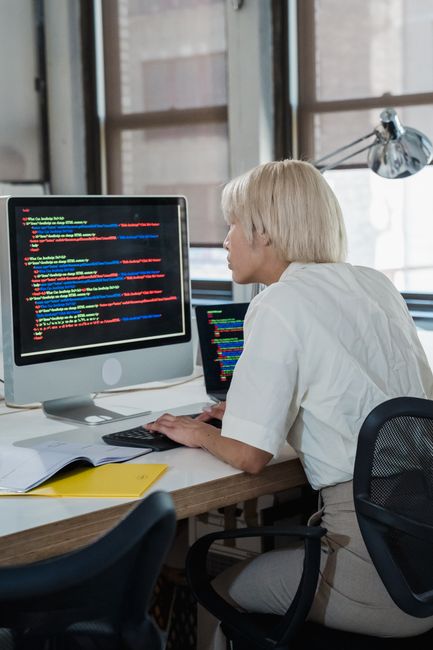 A woman with short blonde hair, dressed in a white shirt, is seated at a desk in a well-lit room with large windows. She is focused on a desktop computer displaying colorful lines of code on the scree